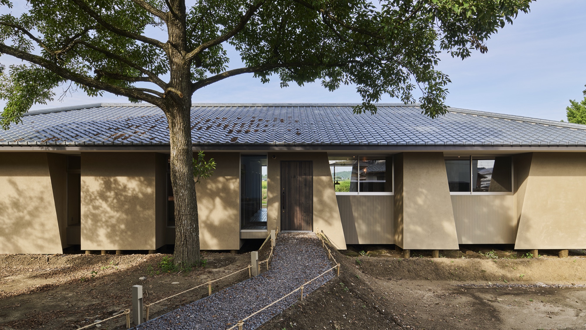 Né seen in daylight — earthen walls in the colour of the ground, a tiled roof, an old tree, the rice fields beyond. / 昼のNé。大地と同じ色の土壁、瓦屋根、一本の古木、その向こうに田。