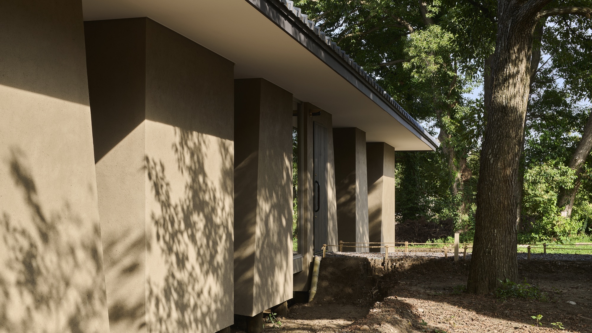 Earthen-walled colonnade under the deep eaves, dappled with the shadows of leaves. / 深い軒下の土壁の列柱に、木漏れ日の影が落ちる。