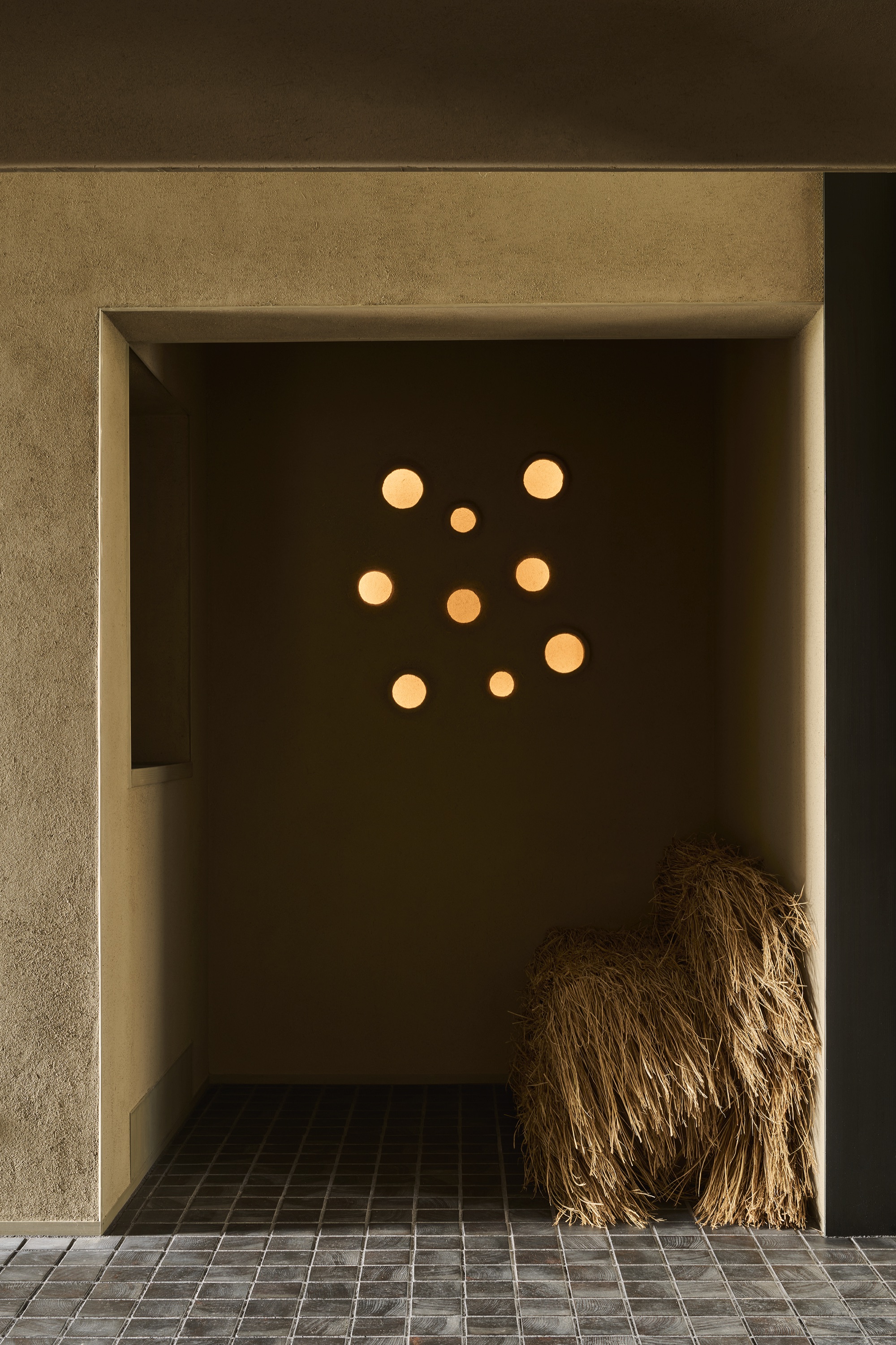A dim, meditative alcove — a constellation of round lights on an earthen wall, a vessel of straw on dark tile. / 薄暗く瞑想的な土間。土壁に円く並ぶ灯り、暗いタイルの上に藁の器。