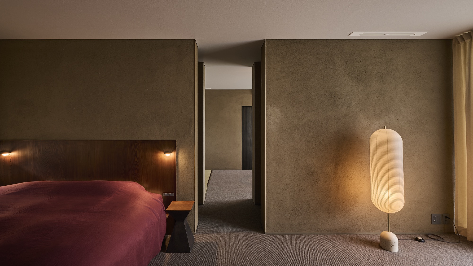 A guest room — a deep red bed, earthen walls, a paper lantern casting soft light. / 客室。深い紅の寝具、土の壁、和紙の灯りがやわらかく照らす。