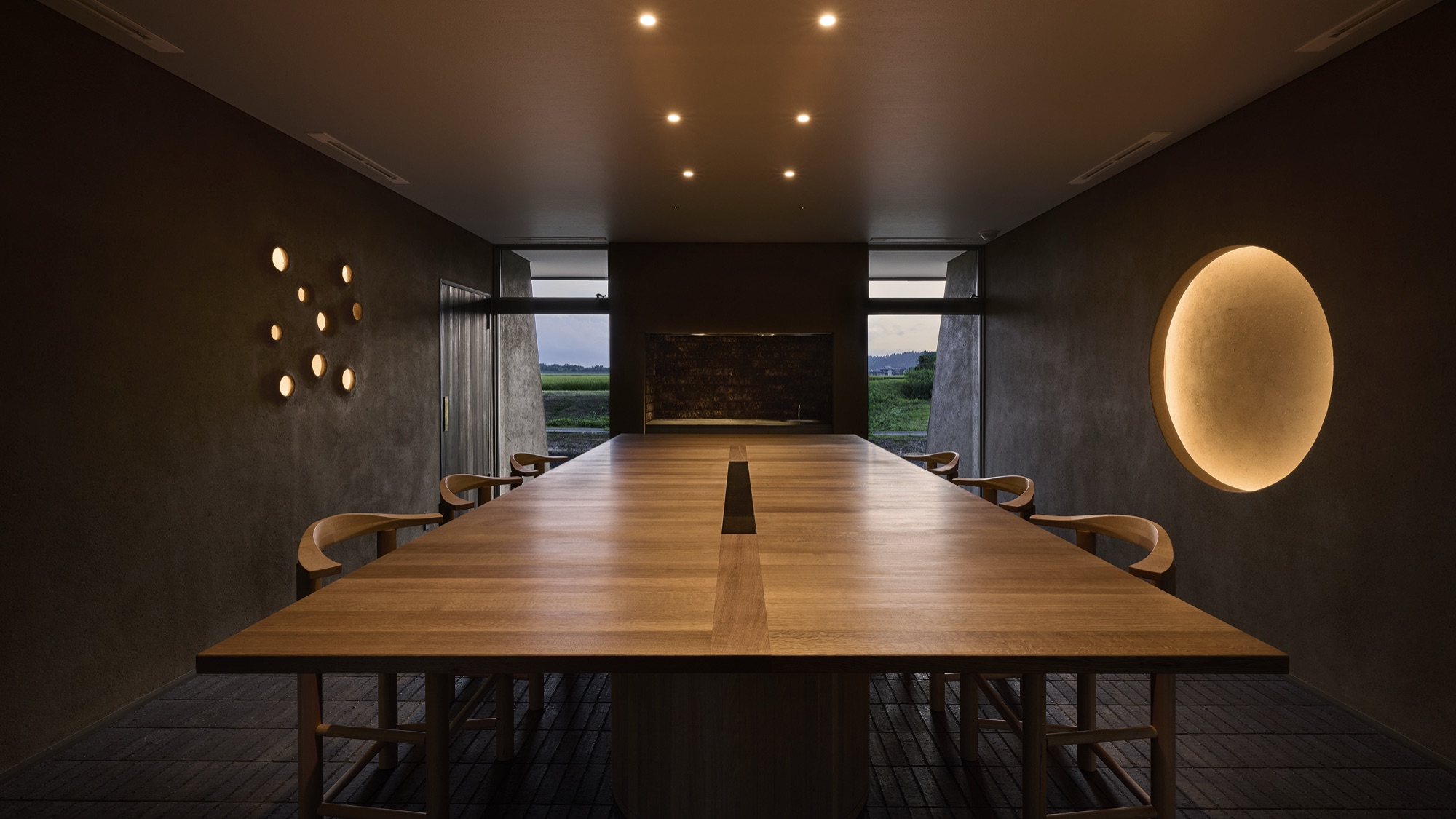 The long single table of solid timber, a moon of light on the wall, the fields visible beyond the windows. / 一枚板の長いテーブル、壁にともる月のような灯り、窓の向こうに田。