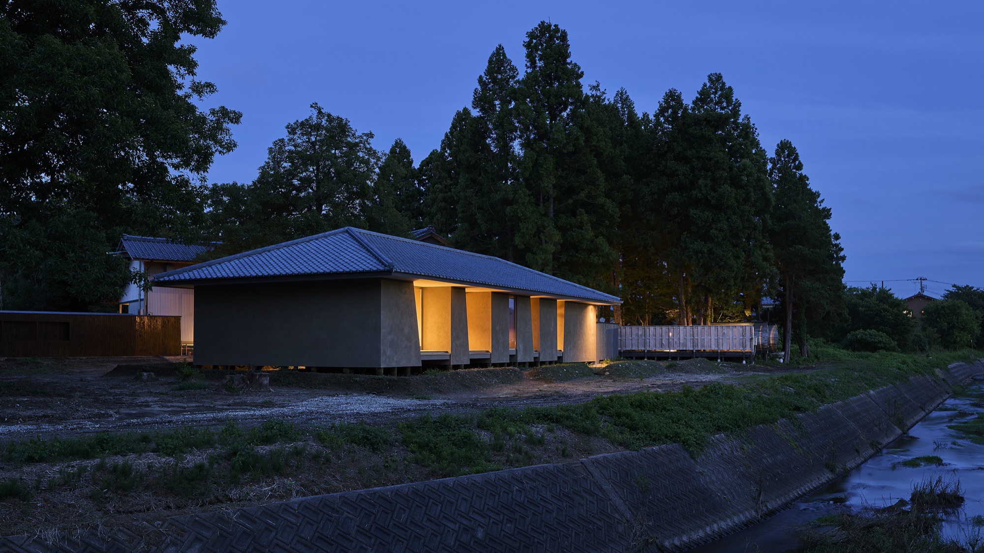 Né at blue hour — a single lit form held within the wide landscape of Shibata, Niigata, tall cedars behind and a river alongside. / ブルーアワーのNé。新発田の広い風景に、灯りをともしたひとつの姿。背後に杉木立、傍らに川。
