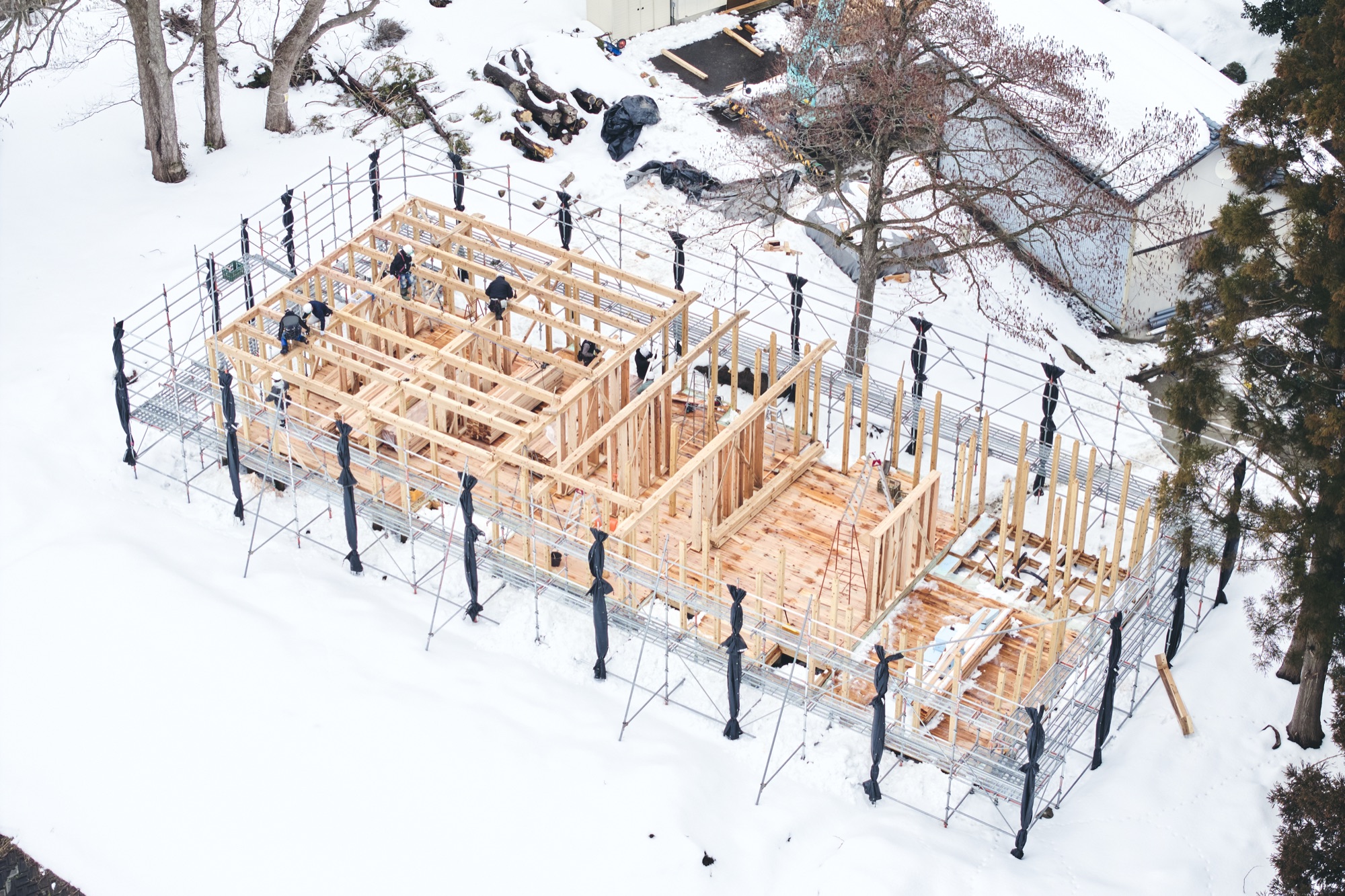 The timber frame of Né rising in deep snow, scaffolding around it, raised by local hands in the depth of winter. / 深い雪の中、足場に囲まれて立ち上がるNéの木組み。真冬に、土地の人の手で。